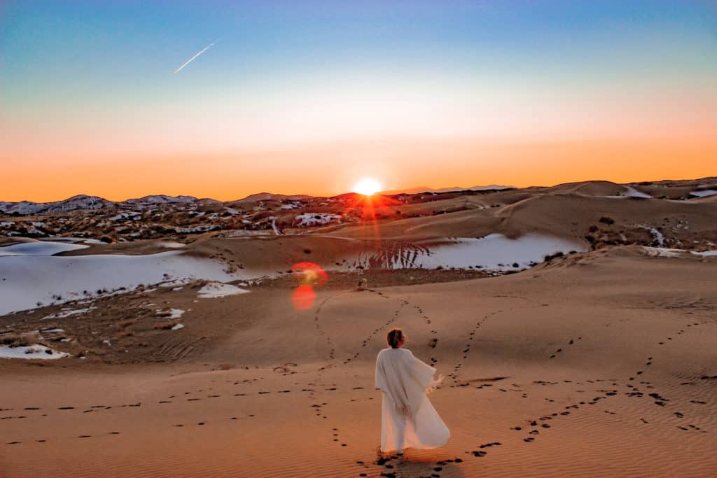 Little Sahara Recreational Area in Utah. Large expanse of sand dunes with patches of snow. An orange and yellow sunset off the horizon. With a dark blue sky beginning to turn to dusk. Little Sahara sand dunes are a great day trip from Salt Lake City full of adventure.