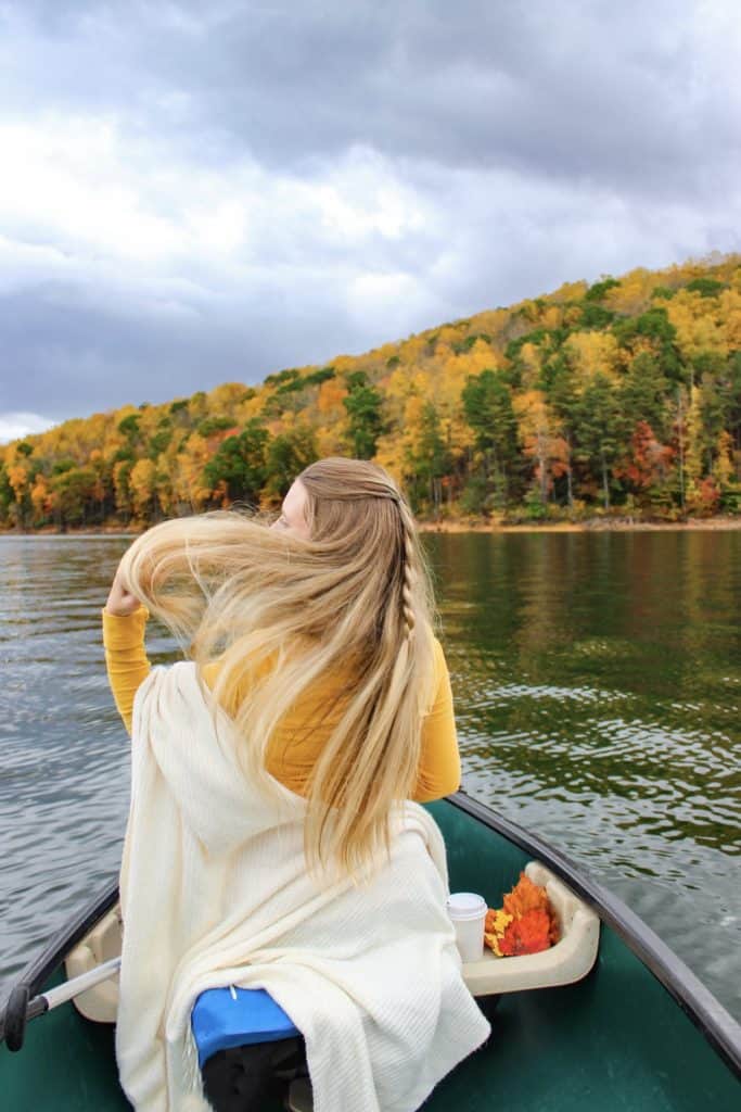 A girl with long blonde hair sitting in a canoe against the backdrop of a gorgeous fall hillside. The changing leaves are yellow orange and red. Canoeing is the perfect fall activity for a day trip. 