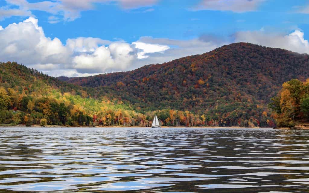 A calm lake with a single sail boat. The surrounding mountains are covered in trees with leaves of every color for fall! Lake Watauga in fall is a sigh to see and lake Watauga is the perfect day trip from Asheville North Carolina. 