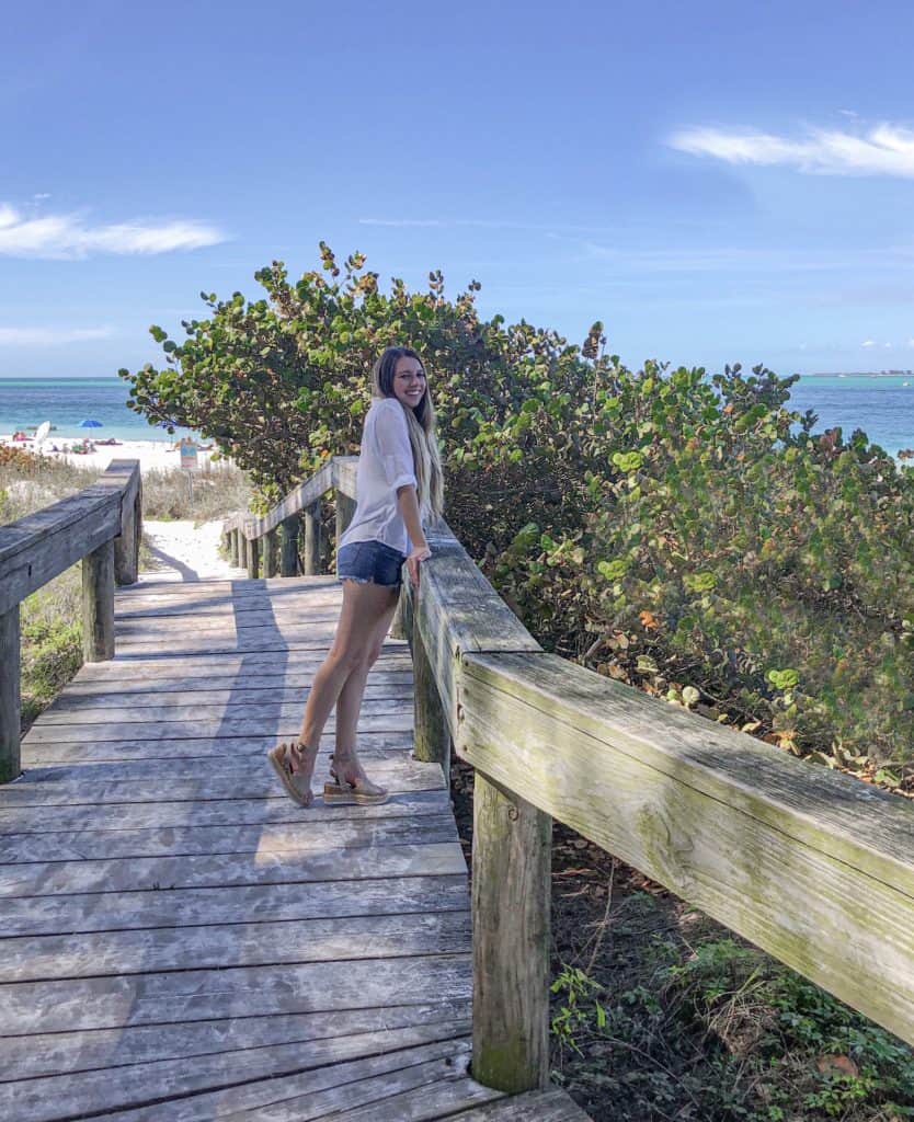 Bright sunny day at Bean Beach on Anna Maria Island, Florida. Girl in summer outfit is on a board walk heading to the white sand beach. The blue water can be seen in the distance.