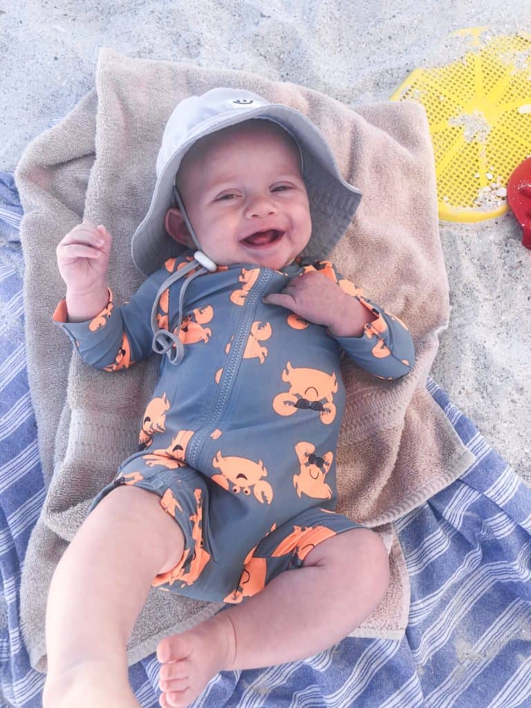 Happy 3 month old baby boy at the beach laying on a beach towel. Every new mom wonders what to bring to the beach with a baby. These are my tried and true baby beach essentials to make every family beach vacation a happy one. From kid friendly sunscreen to infant swimsuits this post talks about it all!