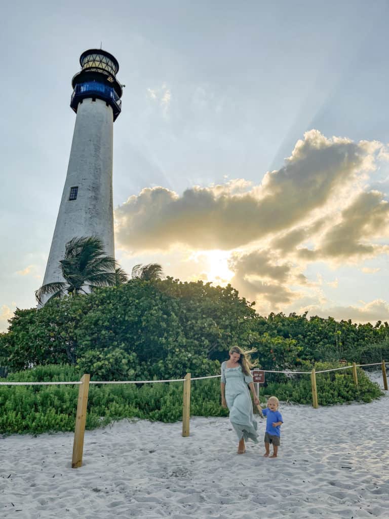 A mother and her 16 month old toddler enjoying sunset on the beach in front of a light house. 