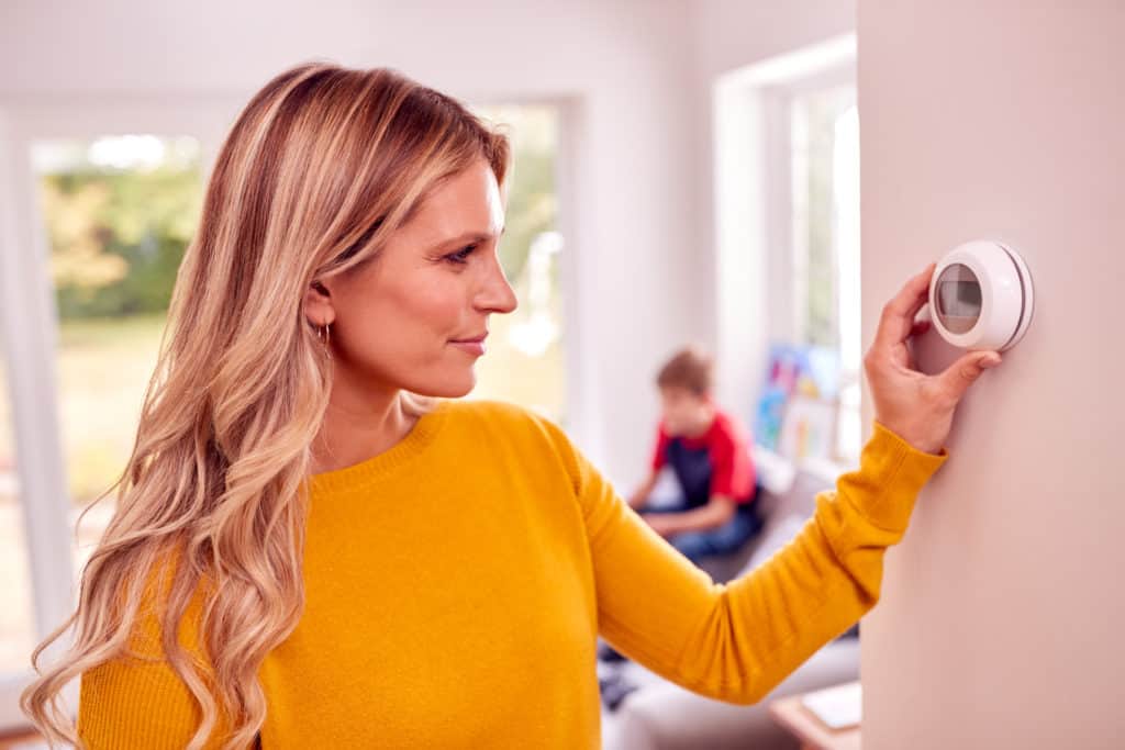 A woman adjusting the thermostat just before leaving on vacation. A smart thermostat allows you to control your homes temperature from anywhere in the world. A crucial step on the leaving home checklist is to raise or lower the temperature of your house to conserve energy.