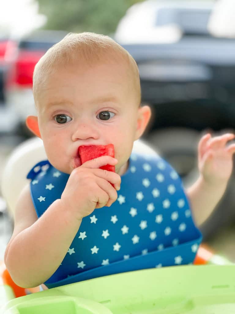 A baby in a silicone bib eating watermelon. Silicone bibs can go anywhere and keep meal time messes to a minimum making them a very important baby travel essential.