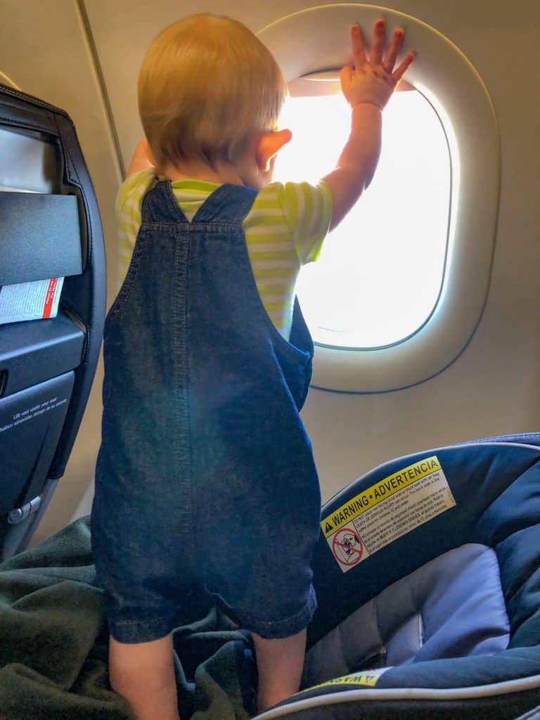 A baby looking out an airplane window while standing in his carseat.