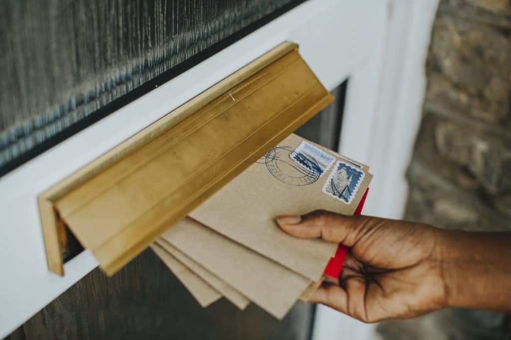 A hand putting a pile of mail through the mail slot of a door. If you don't put your mail on hold before vacation or ask someone to pick it up then the mail piles up which is an easy sign to robbers that the house is empty.