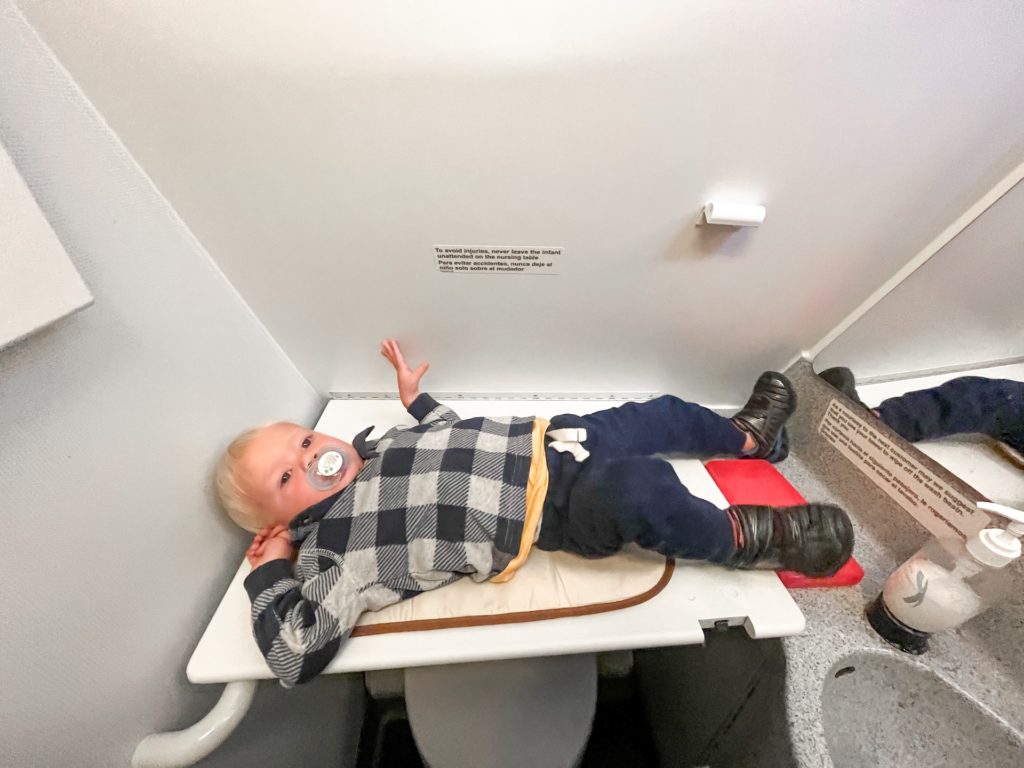 an 18 month old baby boy laying on a changing table on an airplane. Changing diapers on a plane comes with its own unique challenges like turbulence and tight spaces. I am so glad this airplane had a changing table on board.