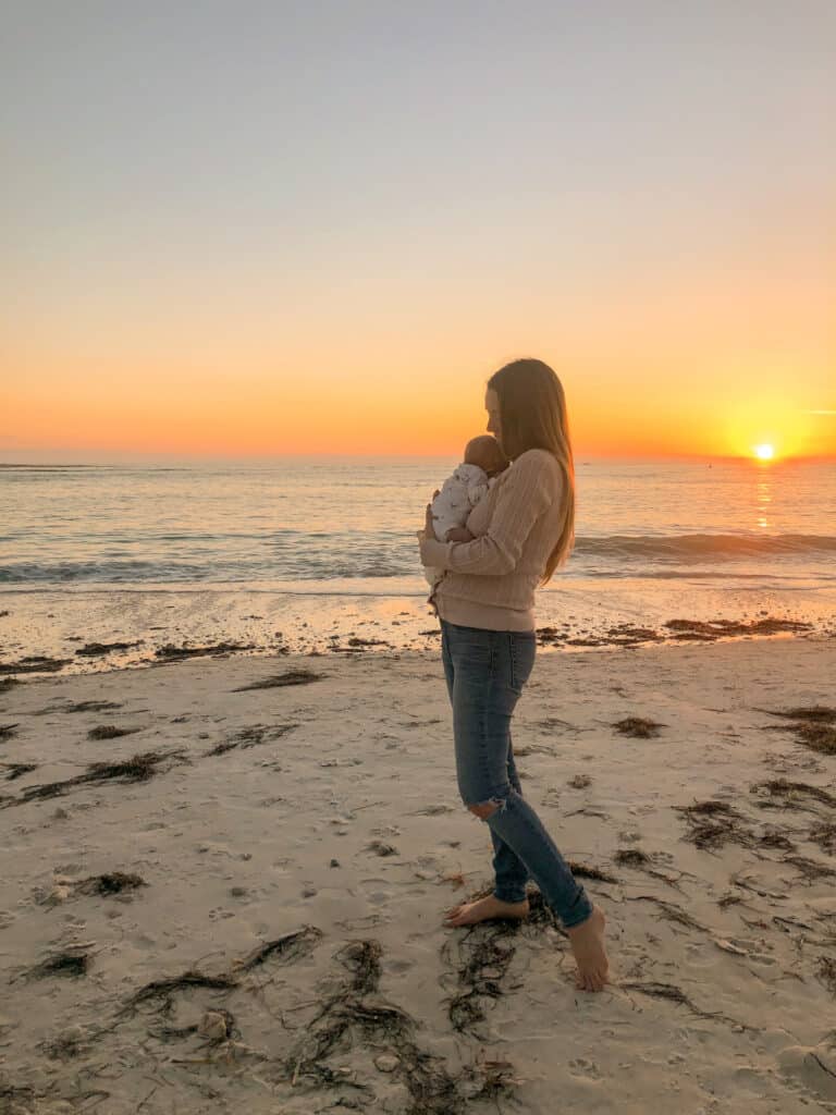 A mother and newborn infant enjoying sunset on the beach. 0-3 months is the worst time to travel with a baby due to their weak immune system. 